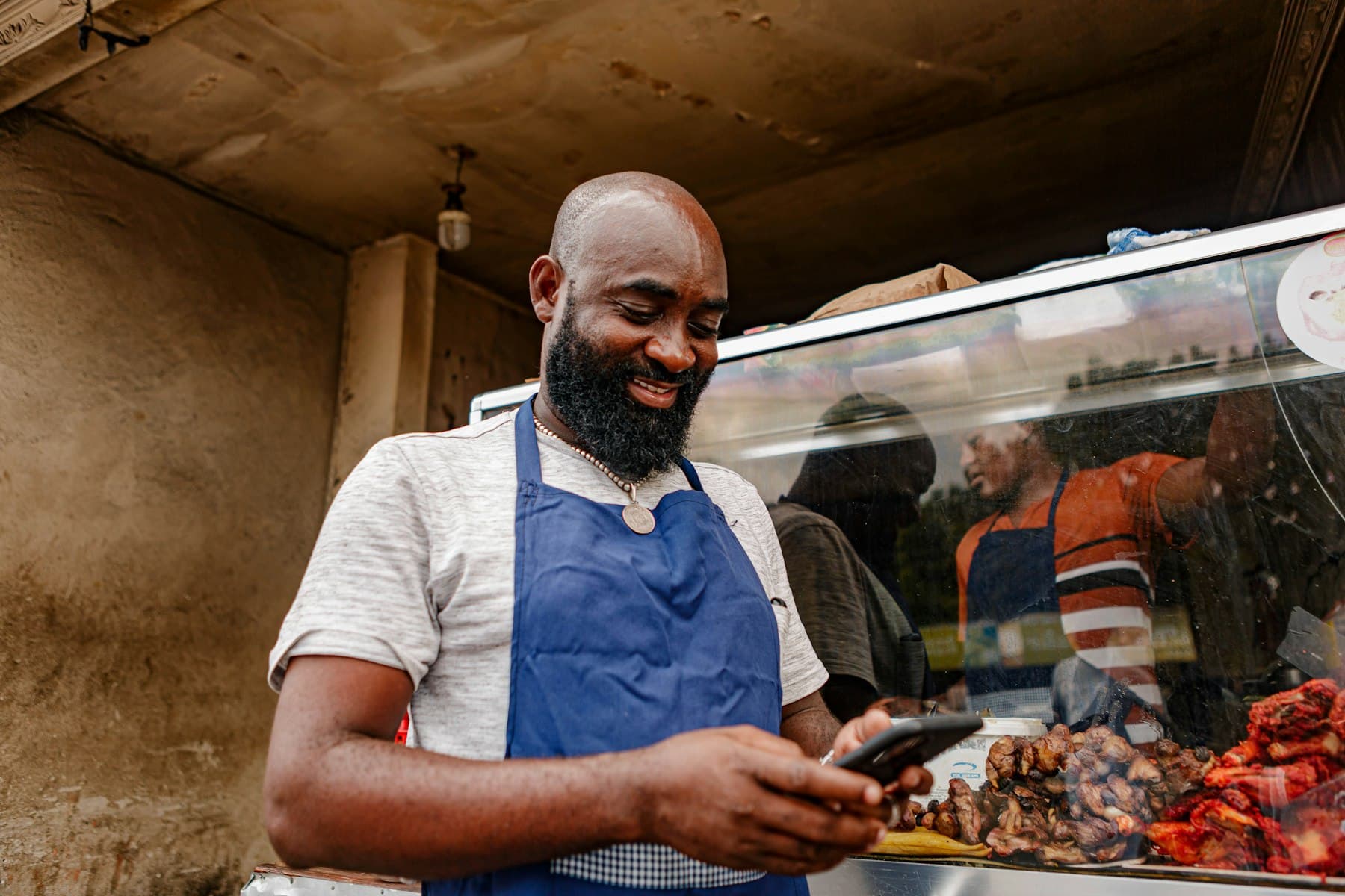 A street food vendor in a blue apron smiles as he checks his phone at his stall.